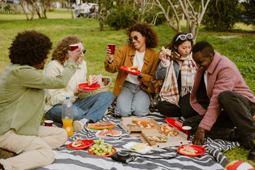 A group of young, diverse friends sit on a blanket enjoying pizza and drinks during a picnic in the park. A concept of friendship, sharing time, and bonding.