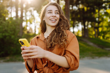 Portrait of cheerful woman with smartphone texting and blogging in sunny park. Young woman relaxing...