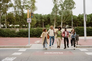 A multinational group of young people walking together across a crosswalk in a sunny urban park...