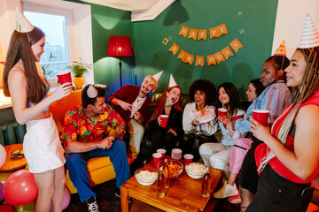 A group of diverse young men and women celebrate a birthday indoors, wearing party hats and holding drinks, with one woman presenting a birthday cake and snacks on a table in a decorated living room.