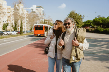 A middle-aged White man and woman walk together near a bus stop in the city, wearing casual sweaters and sunglasses, enjoying a sunny day on the sidewalk.