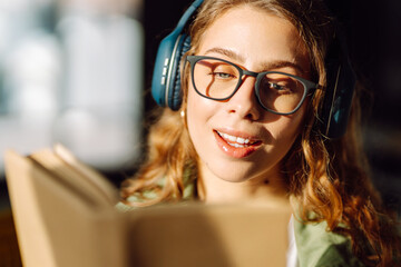 Close-up of a young woman with wireless headphones reading a book in a cozy room. Happy woman...