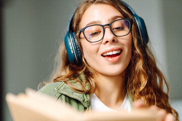 Close-up of a young woman with wireless headphones reading a book in a cozy room. Happy woman...