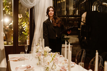 A young White female wedding planner stands next to a set table in a restaurant, checking it out. A...