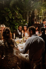 Obraz premium The wedding guests enjoy a candlelit dinner at an elegant indoor reception as the bride and groom sit among them, surrounded by lush greenery and ambient lighting.