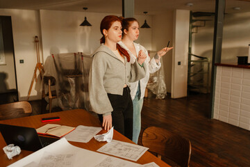 Two women stand beside a work table covered in architectural plans, deep in discussion as they point and gesture in a warmly lit cafe. Concept of teamwork and shared vision in the design process.
