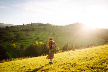 Young female hiker with a hat and a backpack enjoying the mountain landscape at sunset. Beautiful woman feels freedom standing on lush green hills. Hiking, adventure, freedom concept.