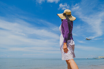a woman wearing a straw hat near the beach look up to the sky when the airplane passing by..