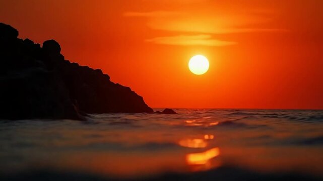 A red burning sunset over the sea with rocky volcanic cliff. Abstract nature summer ocean sea background. Small waves on golden warm water surface with bokeh lights from sun.