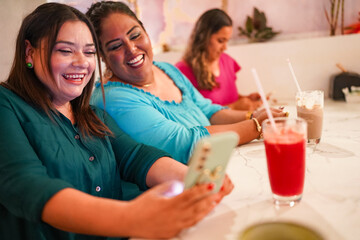 Latin women sharing funny content on smartphone while drinking juice at cafe