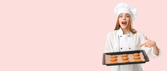 Young female chef pointing at cookies on baking tray against pink background
