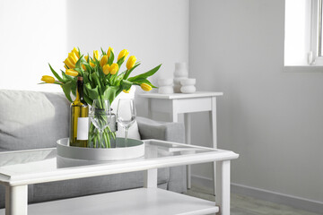 Beautiful vase of yellow tulips with bottle of wine and glasses on coffee table in living room
