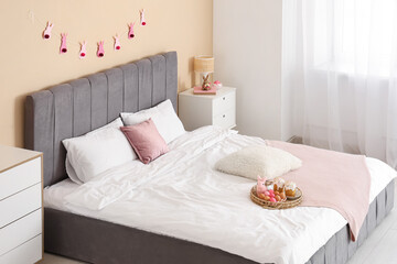Wicker tray with Easter cakes, painted eggs and bunny on bed in festive bedroom
