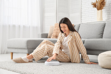 Young woman in pajamas turning robot vacuum cleaner on carpet at home