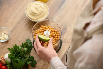 Young woman adding lime juice into blender with chickpeas for hummus on table in kitchen