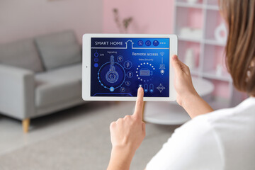 Young woman using tablet with smart home security system control panel in room, back view