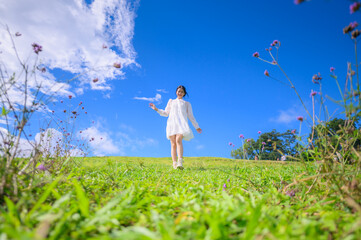 A happy young woman in a flowing white dress strolls through a vibrant field of flowers under a bright blue sky with white clouds