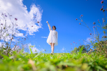 A happy young woman in a flowing white dress strolls through a vibrant field of flowers under a bright blue sky with white clouds