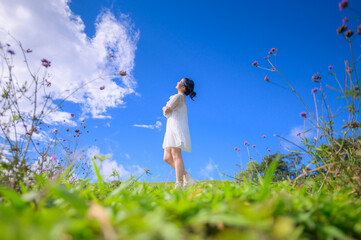 A happy young woman in a flowing white dress strolls through a vibrant field of flowers under a bright blue sky with white clouds