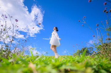 A happy young woman in a flowing white dress strolls through a vibrant field of flowers under a bright blue sky with white clouds