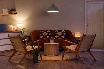 Interior of stylish living room with brown sofa, armchairs, glowing lamps and surfboard at evening