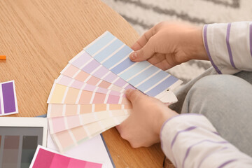 Young woman with paint palette swatches choosing lilac color near table at home, closeup