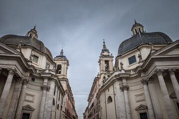 Fototapeta premium Low-angle view of Rome's twin Baroque churches - Santa Maria dei Miracoli (left) and Santa Maria in Montesanto - framing Via del corso at Piazza del Popolo beneath an overcast sky in rome, italy.