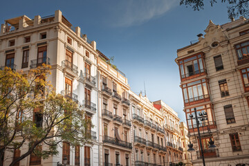 Apartment blocks in Ciutat Vella show wooden shutters, wrought-iron balconies and early-20th-century facades framed by treetops and clear sky, illustrating Mediterranean residential real-estate charm.