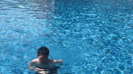 Asian boy swimming in a blue-tiled pool with ripple patterns and sunlit water. Solo child swimmer performing strokes on a bright summer day. Fun, health, and motion in outdoor leisure setting.