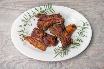 Barbecued pork ribs on a white plate with rosemary: a delicious home-cooked meal on a rustic wooden table.