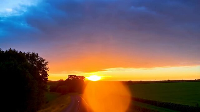 Classic vintage car driving down a rural road at sunset reflecting vibrant orange and purple colors onto the windshield, with green fields on either side.