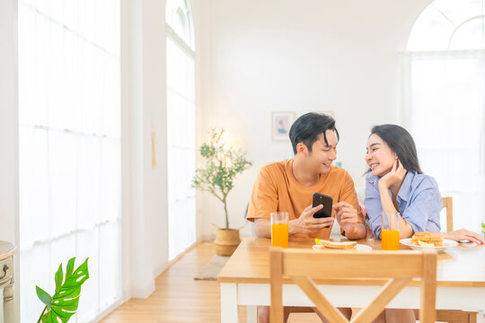 Happy Asian family couple using smartphone with internet online shopping or electronic banking at home. Husband and wife enjoy breakfast together. People using AI on mobile phone helping smart life.