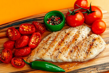 A grilled white fish fillet seasoned with black pepper and spices is served on a wooden board with fresh and roasted cherry tomatoes, a green chili pepper, and a bowl of mixed peppercorns.