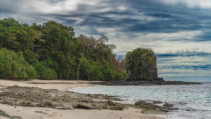 A secluded beach on a tropical island, without people. A scattering of boulders on a sandy beach, thickets of green vegetation. A calm turquoise ocean. A picturesque rock against a  blue sky, clouds. 
