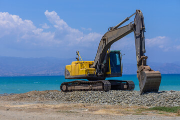 A yellow excavator levels the sand on a beach in Cyprus. Construction or maintenance work under clear blue sky near the Mediterranean Sea.