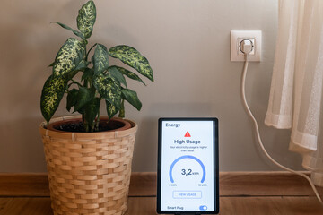 Smart home device displaying a high energy consumption alert on a digital tablet beside a plant and wall plug, emphasizing energy saving and efficient management