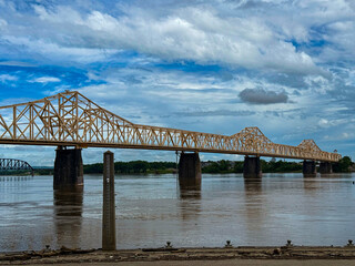 Obraz premium Yellow Truss Bridge Spanning a River Under a Cloudy Blue Sky