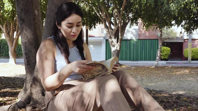 Mujer joven leyendo un libro sentada bajo un &aacute;rbol.