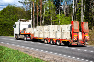 A white truck with a lowboy semitrailer transports concrete products along a country road against a forest background. Oversized cargo. Copy space for text