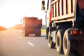 Orange road dump trucks drive along the highway after delivering asphalt against the backdrop of sunset in summer, road repair. Copy space for text, industry