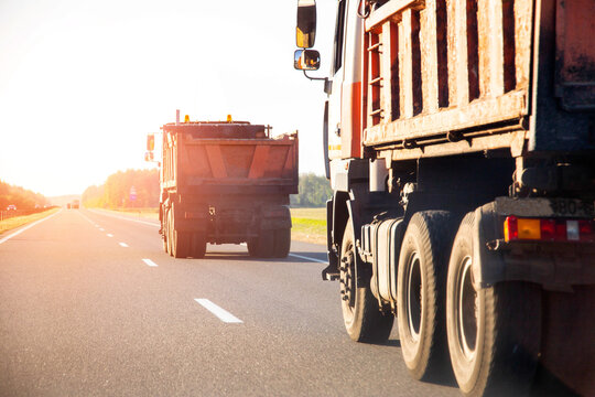 Orange road dump trucks drive along the highway after delivering asphalt against the backdrop of sunset in summer, road repair. Copy space for text, industry
