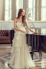 Beautiful slim young woman in white luxurious wedding dress. Preparation for the wedding, romantic morning of the bride. Girl with beautiful long hair stands at the piano.