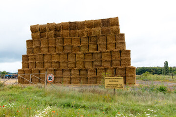In a tranquil field in France, a tall structure of straw bales stands stacked against a backdrop of overcast clouds. The area is marked for public safety.