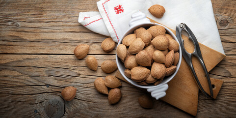 Whole almonds in shell in white ceramic bowl with old nutcracker on rustic wooden background, top view, space for text.