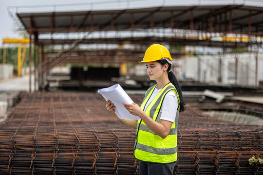 Female civil engineer or architecture is using a blueprint plan to check for the progress of construction. Worker with safety suit and walkie talkie is inspecting the factory production system outdoor