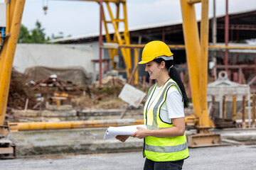 Female civil engineer or architecture is using a blueprint plan to check for the progress of construction. Worker with safety suit and walkie talkie is inspecting the factory production system outdoor