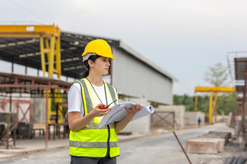 Female civil engineer or architecture is using a blueprint plan to check for the progress of construction. Worker with safety suit and walkie talkie is inspecting the factory production system outdoor