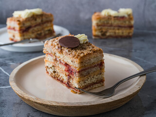 Piece of layered cake with cream and berry filling on beige plate, two more pieces blurred in the background. Marble surface. Cozy and elegant dessert scene.