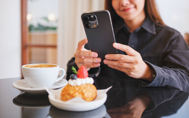 Closeup of a woman holding and using mobile phone with coffee cup and cake on the table in cafe
