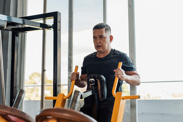 A determined Southeast Asian man in his 50s performs seated rows on a plate-loaded chest supported row machine at an open-air gym. Training back.
